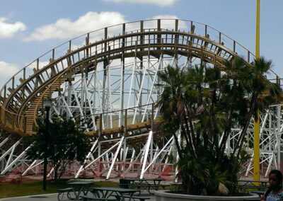 A wooden roller coaster with white supports under a blue sky, surrounded by trees and picnic tables.