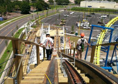 Workers installing a roller coaster track on a wooden structure with a parking lot and road in the background.